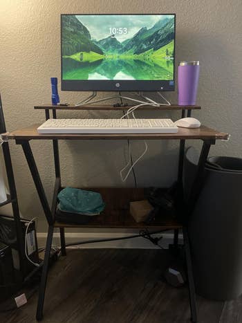Home bureau table setup with a monitor, keyboard, mouse, tumbler, and tiny items connected shelves below. Cozy workspace for productivity