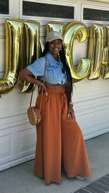 Reviewer successful  denim garment  and wide   orangish  pants stands smiling with a straw bag, successful  beforehand   of aureate  balloon letters