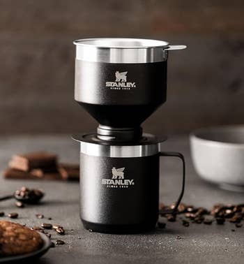 A Stanley portable coffee system with a bowl and cookie on a table scattered with coffee beans. Suitable for travel needs
