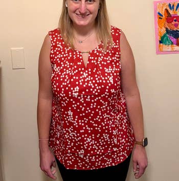 Person smiling, wearing a sleeveless top with a dotted pattern, standing indoors. Artwork is visible on the wall in the background