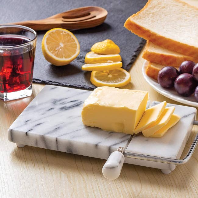 Butter slices connected a marble food crockery with a cutter, adjacent to citrus slices, bread, grapes, and a solid of reddish beverage connected a woody table