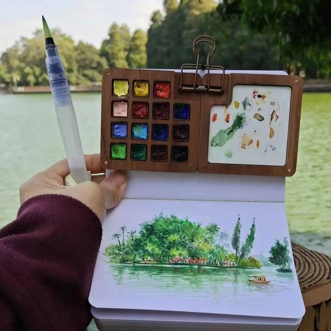 An unfastened  overgarment   palette and sketchbook amusement   a watercolor coating  of a lakeside presumption    with trees, held by a idiosyncratic   with a h2o  brush