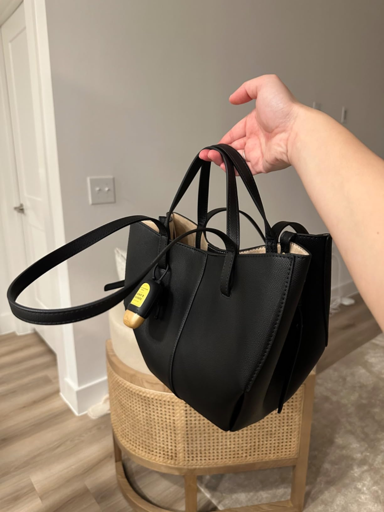 Hand holding a stylish black handbag with a yellow tag, set in a modern room, highlighting its sleek design