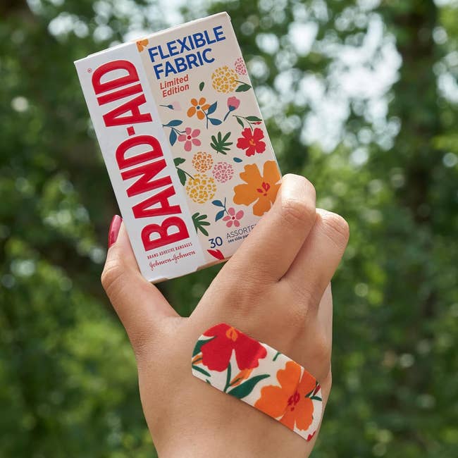 Hand holding floral-design Band-Aid container and wearing matching bandage, against a leafy background