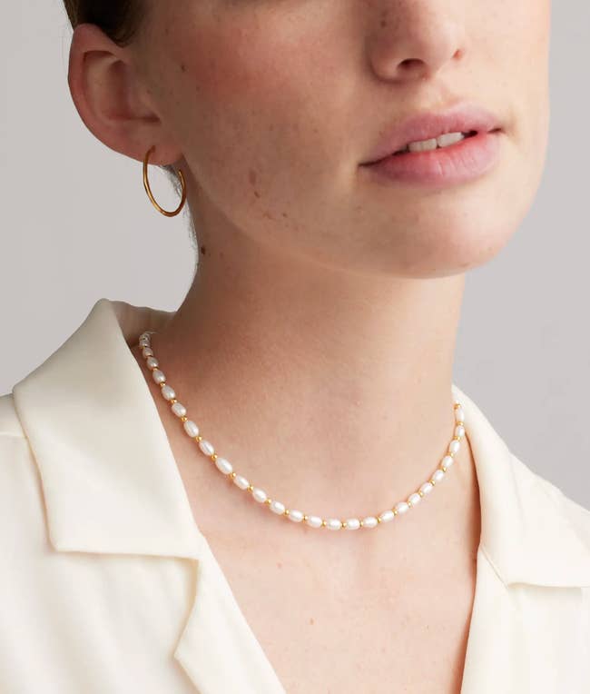 Person wearing pearl necklace, golden  hoop earring, and achromatic  collared blouse, showcasing elemental  and elegant accessories