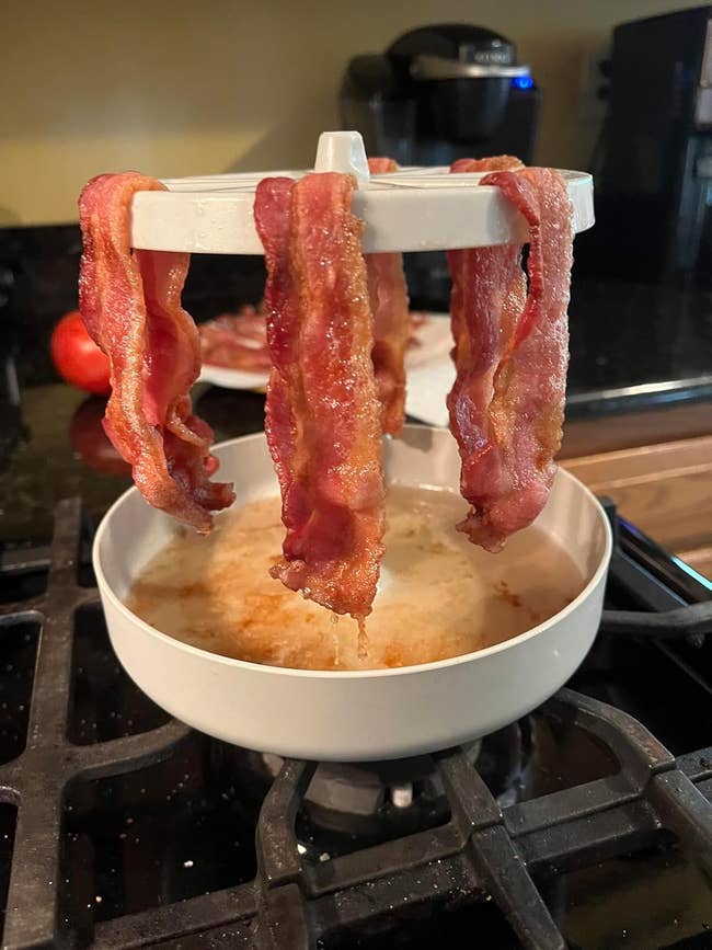 Bacon slices hang on a rack over a bowl on a stovetop, draining grease