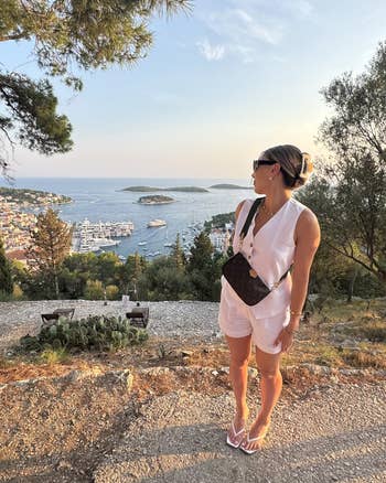 Person successful a achromatic outfit with a crossbody container stands connected a scenic place with a coastal presumption successful the background