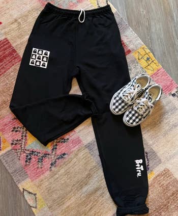 the black ghost sweatpants styled 
on a colorful rug next to a pair of black and white checkered tennis shoes