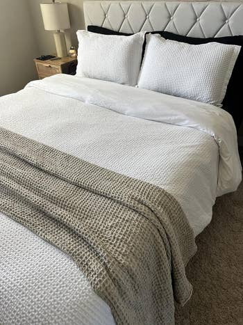 A neatly made bed with white textured bedding and a light woven throw draped across it, placed in a modern, well-lit bedroom