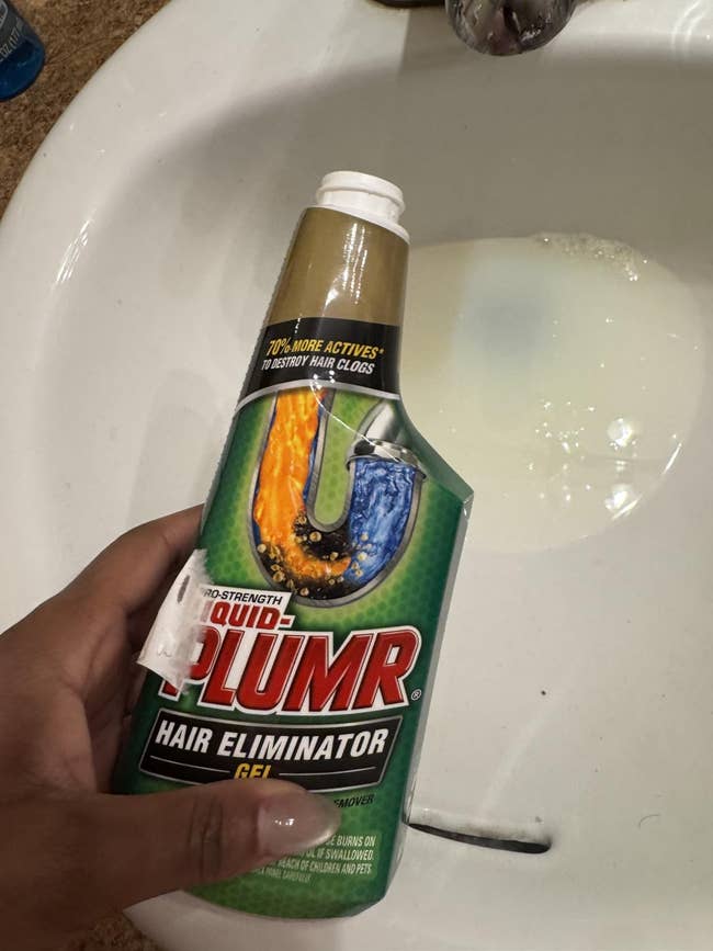 A hand holds a bottle of Liquid Plumr Hair Eliminator Gel above a bathroom sink with water in it
