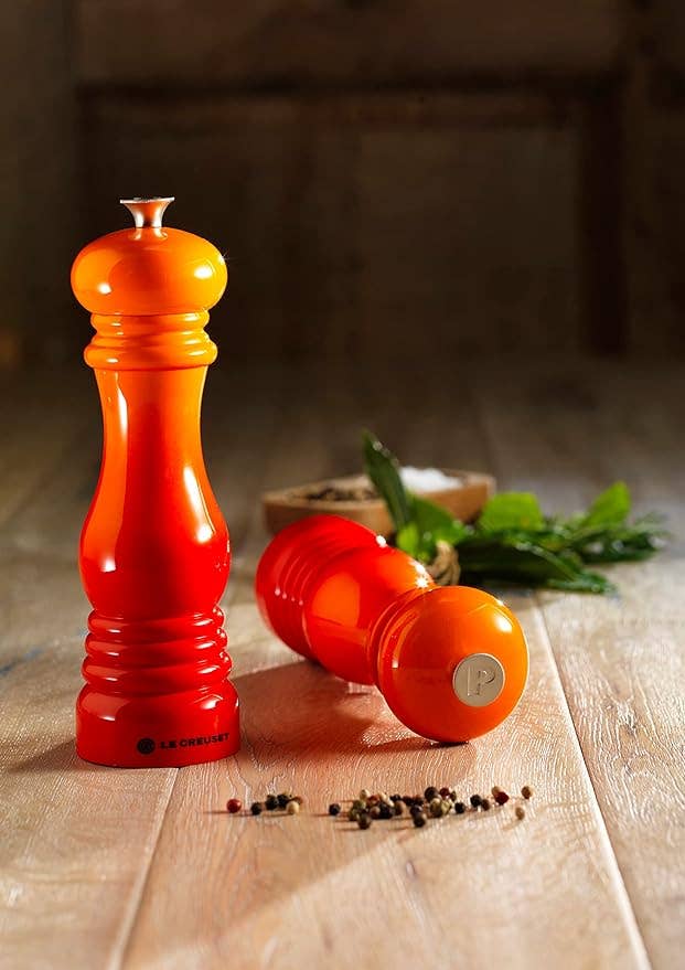 Two orange and red gradient Le Creuset pepper mills on a wooden surface, one standing upright and the other lying on its side