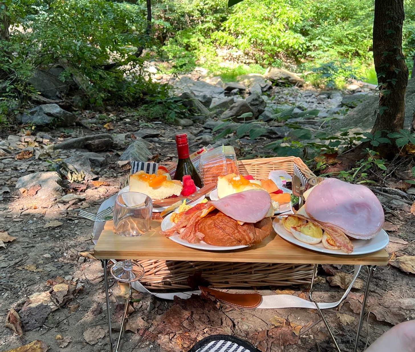 Pengaturan piknik di atas meja kecil di tempat terbuka dengan piring croissant, ham, telur, irisan jeruk, sebotol anggur, dan gelas anggur