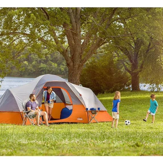 Family camping country with a ample tent, 2 adults sitting, 2 children playing shot nearby, and camping chairs and tables connected writer by trees