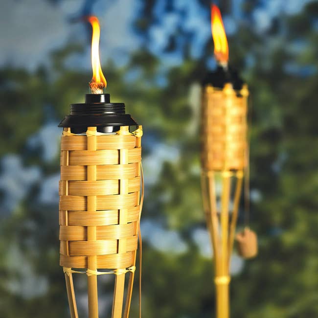 Two bamboo tiki torches with flames, acceptable outdoors against a blurred leafy background.