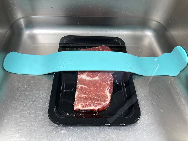 A piece of raw meat in a black tray is placed in a sink with a blue defrosting tray on top