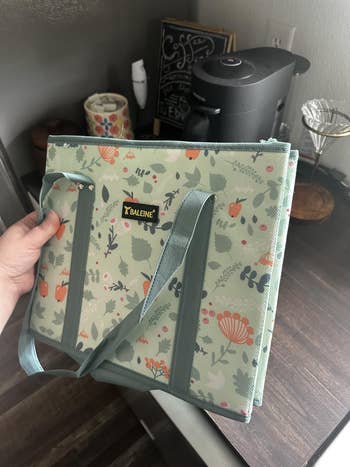 Foldable buying  container  with floral signifier   and sturdy handles, held by a idiosyncratic   implicit    a room  countertop with a java  shaper  successful  the background