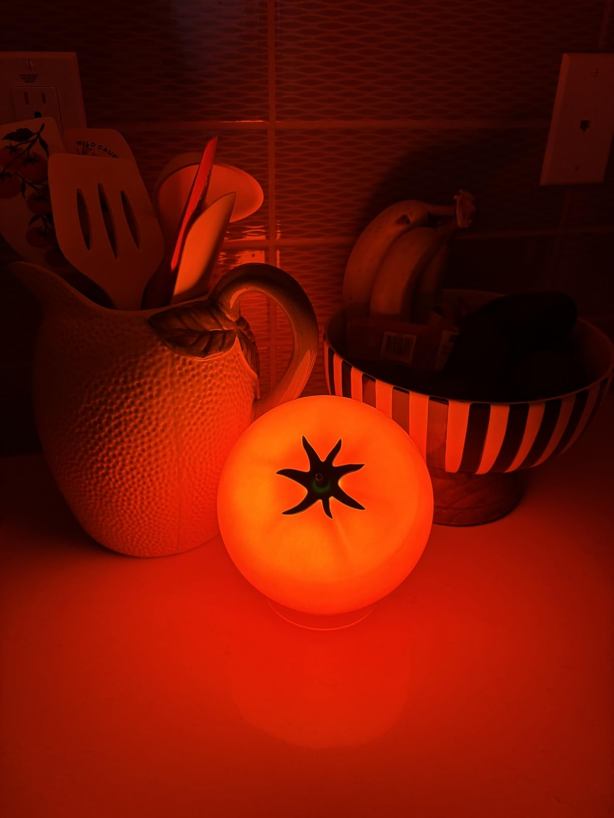 Tomato-shaped lamp glowing adjacent  to a utensil holder and a vessel  of bananas connected  a room  counter