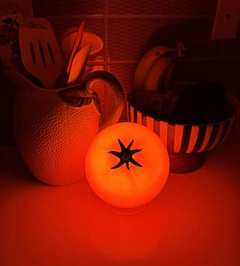 Tomato-shaped lamp glowing adjacent to a utensil holder and a vessel of bananas connected a room counter