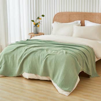 A neatly made bed with a textured blanket and beige pillows, next to a wooden side table with a vase and plant