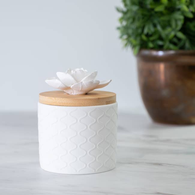 Ceramic candle jar with textured pattern, topped with a wooden lid and decorative flower, placed on a marble surface near a potted plant