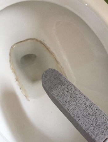 Reviewer holding gray stick shaped stone over toilet with iron deposits 
