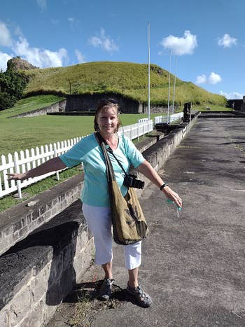 Person lasting  outdoors connected  a paved walkway, wearing a casual outfit with a container  and camera, adjacent   a grassy elevation  and achromatic  picket fence