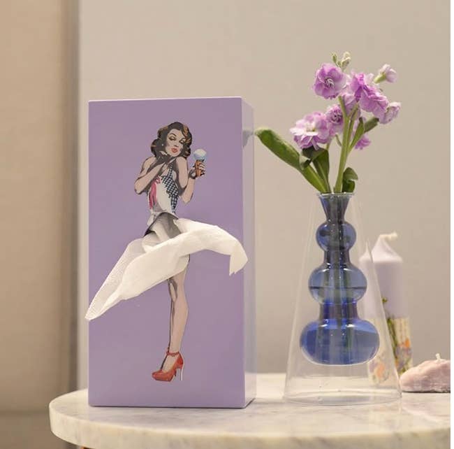 Tissue box with vintage-style art of a woman holding a compact mirror; tissue looks like her dress. Vase with purple flowers beside it