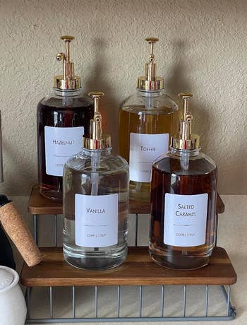 Four glass bottles of coffee syrup on a wire rack labeled Hazelnut, Toffee, Vanilla, and Salted Caramel, with gold pump tops