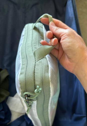 A hand holding a green packing cube with a zipper and handle, inside a suitcase