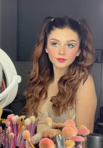 Reviewer with colorful winged eyeliner and two hair buns, surrounded by makeup brushes