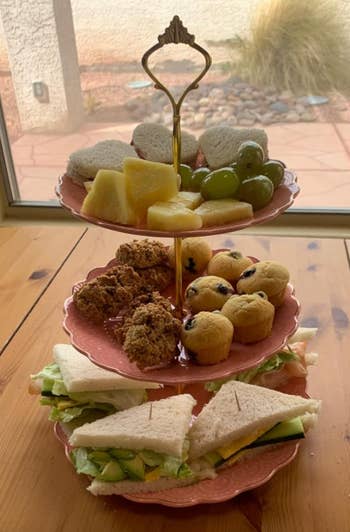 Three-tiered serving tray with sandwiches on the bottom, baked goods in the middle, and fruit on top, displayed indoors