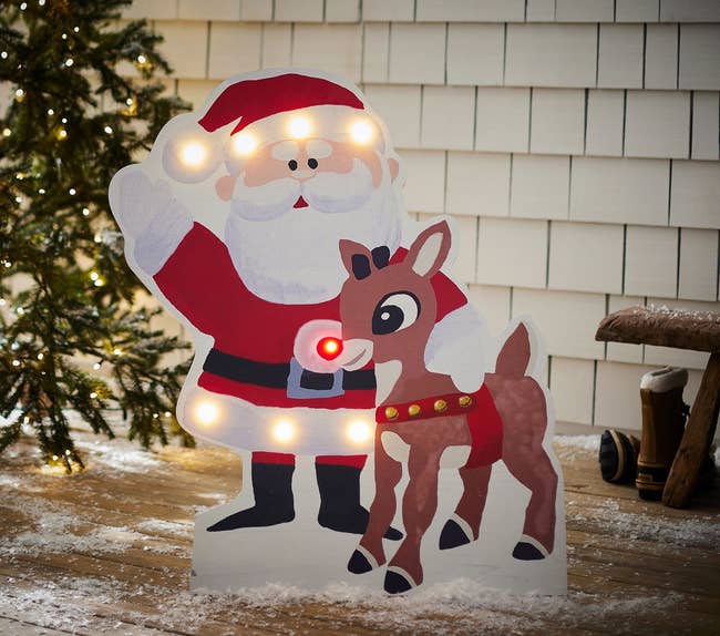 Santa and reindeer cut-out decoration lit with bulbs, set on a snowy porch near a tree, featured in a holiday shopping article