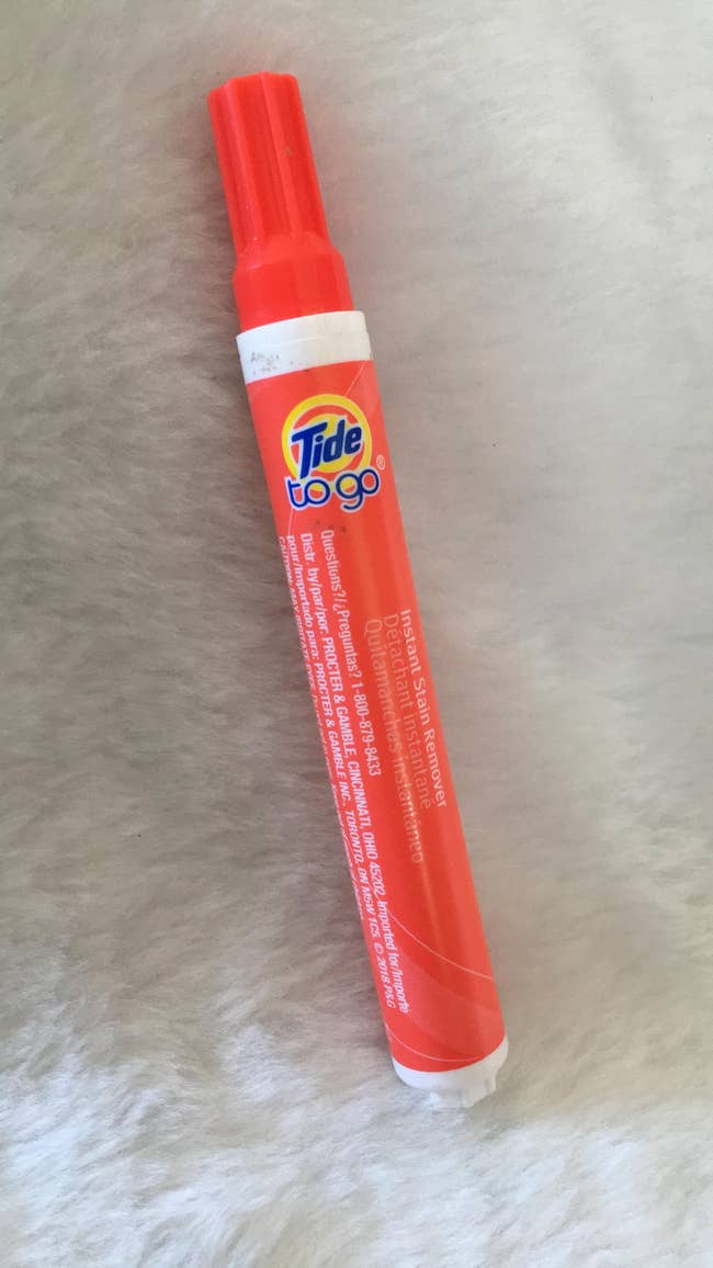 Tide to Go Instant Stain Remover pen, reddish  packaging with cap, laying connected  a soft, light-colored surface. Ideal for speedy  stain removal connected  the go