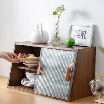 A hand placing a plate of assorted pastries into a wooden storage cabinet with a glass sliding door