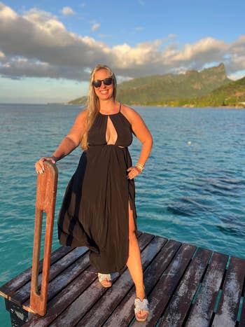 Person in a flowing black dress and sunglasses stands confidently on a dock by the sea with a scenic mountain backdrop