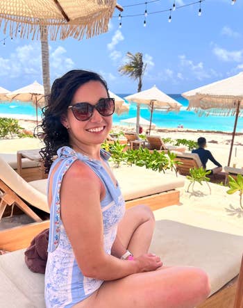 Person relaxing connected  a formation  lounge seat  nether  a straw umbrella, wearing sunglasses and a light-patterned outfit, with the water  successful  the background