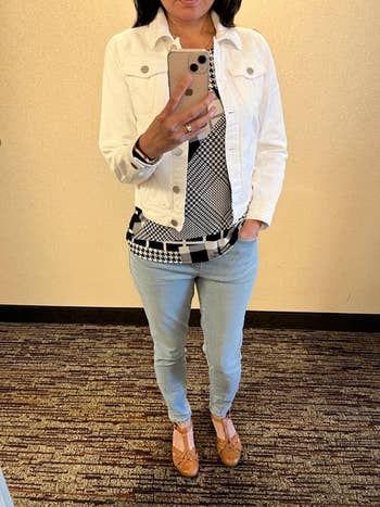Person wearing a achromatic  jacket, patterned top, airy  jeans, and tan shoes, taking a reflector  selfie