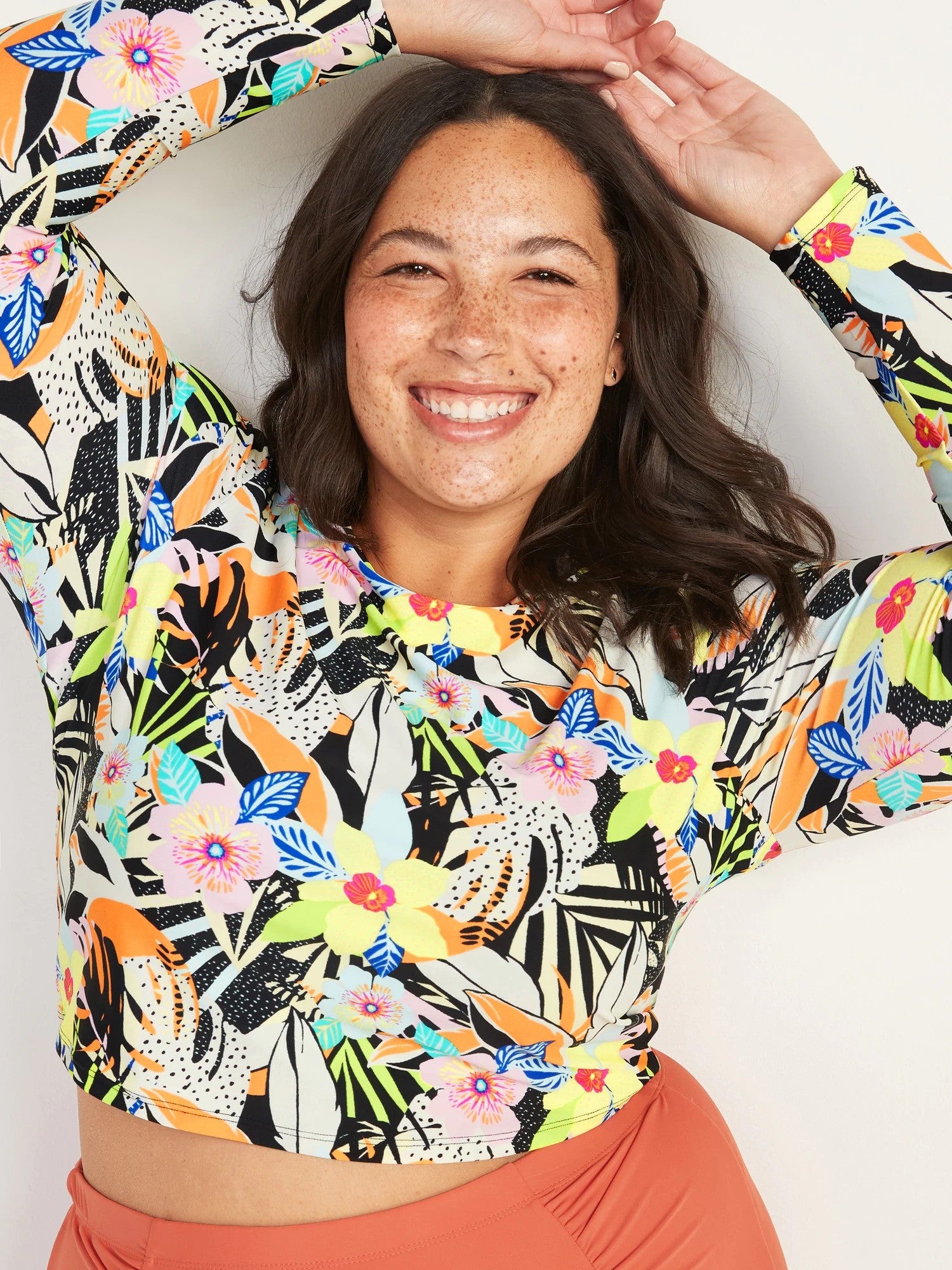 model wearing top with illustrated monstera leaves, hibiscus flowers, palm leaves, and orchid flowers in black, orange, pink, blue, yellow