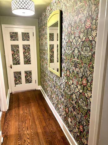Hallway with patterned floral wallpaper, woody floor, a achromatic doorway with solid panels, and a partition mirror