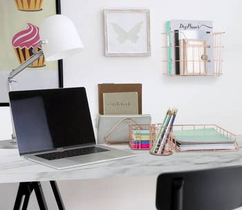 A modern home office desk setup with a laptop, lamp, notebooks, pens, and a wall organizer. Cupcake art and a butterfly print decorate the wall