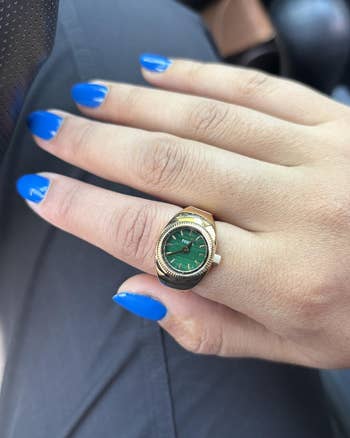 Hand with bluish nails displaying a golden ticker with a greenish face, worn arsenic a ring