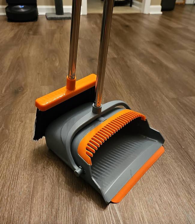 An orange broom with a gray dustpan with slotted edges to clean the bristles of the broom 
