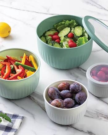 Assorted vegetables and fruits in round, textured storage containers on a marble surface, with a lemon and a checkered cloth nearby