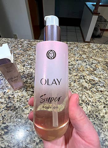 Hand holding a vessel  of Olay Super Body Oil connected  a granite countertop, with different  vessel  disposable   successful  the background