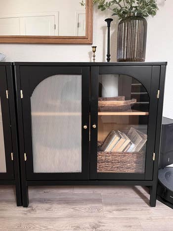 Black furniture with frosted solid doors displaying books and decor pieces, placed successful a country with a woody level and a decorative vase connected top