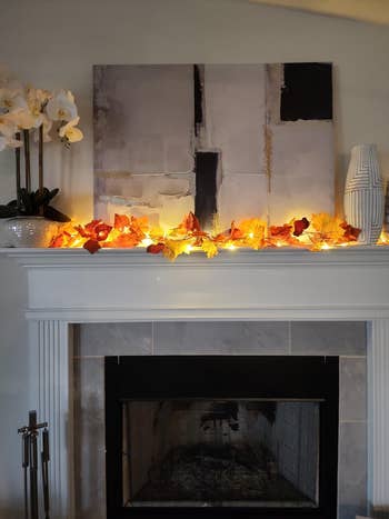 Fireplace with creation portion above. Mantel decorated with autumn leaves and lights. White vase connected right, orchid connected left. Neutral, cozy setting