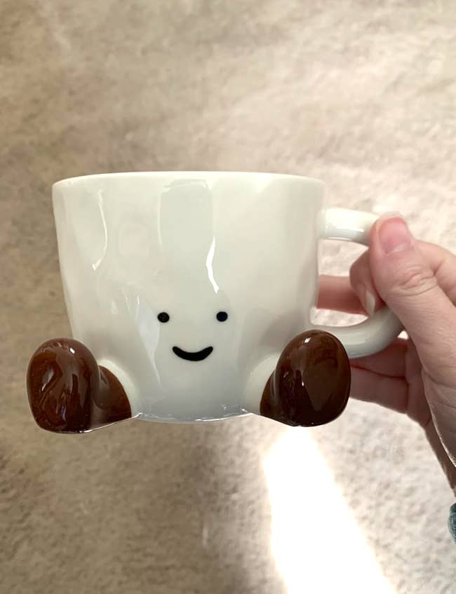 Ceramic mug with a smiling look and brownish feet, held by a hand