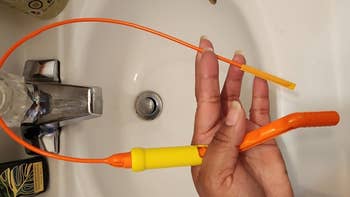 Hand holding a flexible, plastic drain clog remover tool with a barbed end, positioned over a bathroom sink