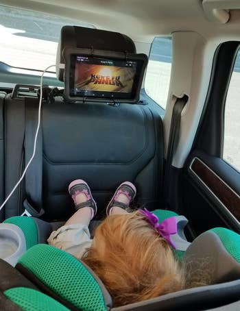a reviewer photo of the mount on a headrest with a child in a rear-facing car seat watching it 