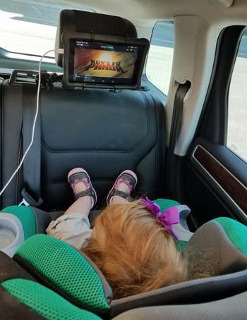 a reviewer photo of the mount on a headrest with a child in a rear-facing car seat watching it 